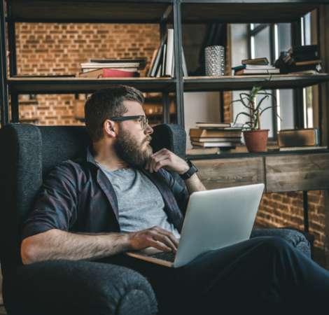 Young bearded man in eyeglasses using laptop while sitting in armchair at home